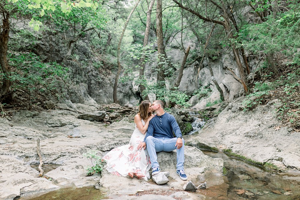 Couple sitting on rocks kissing surrounded by trees and stream